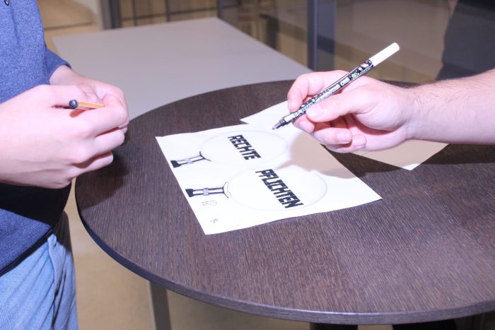 Zwei Lehrlinge stehen vor einem Tisch mit Stiften in der Hand und zeichnen auf einem Blatt.