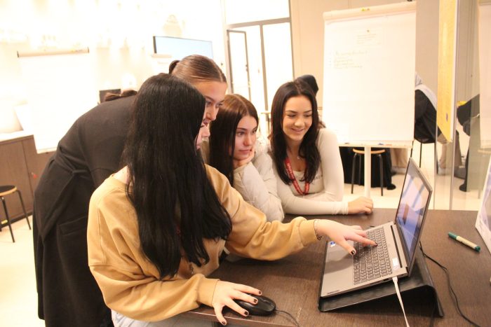 3 Mädchen sitzen, 1 Mädchen seht vor einem Tisch mit einem Laptop und zeigen auf den Bildschirm.
