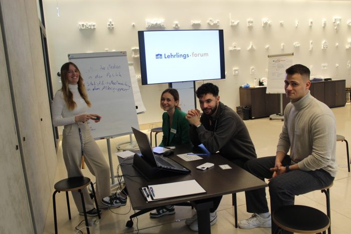 3 Lehrlinge sitzen auf Hocker vor einem Tisch mit einem Laptop und Unterlagen, 1 Lehrling steht vor einem Flipchart.