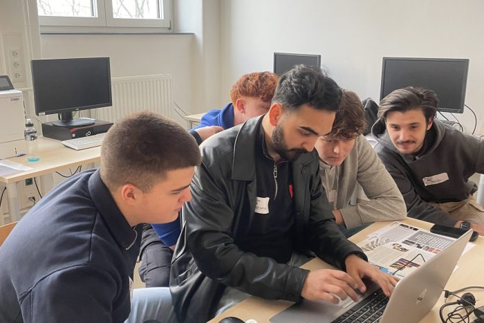 5 Lehrlinge sitzen vor einem Tisch mit einem Laptop mit Unterlagen, 1 Lehrling schreibt.