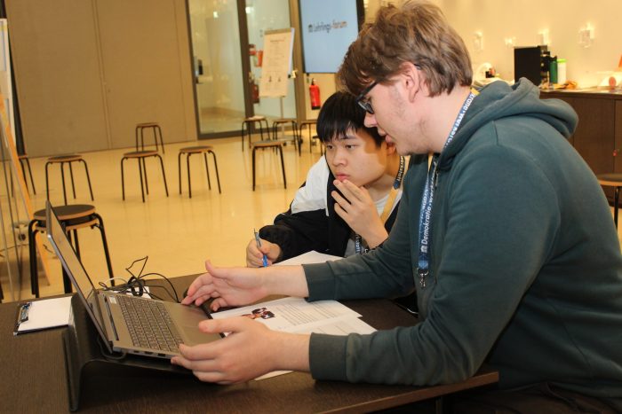 2 Lehrlinge sitzen vor einem Tisch mit einem Laptop und Unterlagen und unterhalten sich.