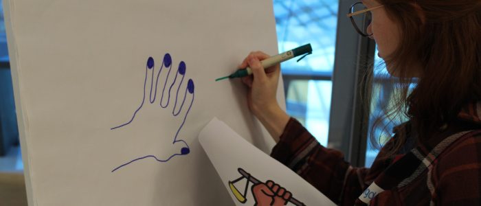 : Ein Lehrling steht vor einem Flipchart mit einem Blatt und Stift in der Hand und zeichnet.