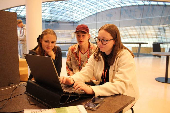 Drei Lehrlinge sitzen vor einem Tisch mit einem Laptop und Unterlagen darauf, ein Lehrling schreibt.