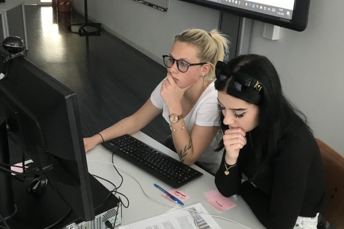 Zwei Lehrlinge sitzen an einem Tisch mit Computer, im Hintergrund ist eine Leinwand zu sehen
