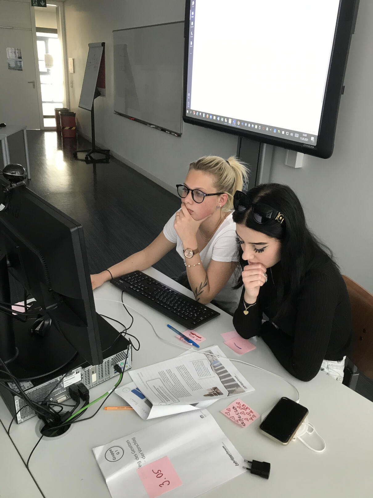 Zwei Lehrlinge sitzen an einem Tisch mit Computer, im Hintergrund ist eine Leinwand zu sehen